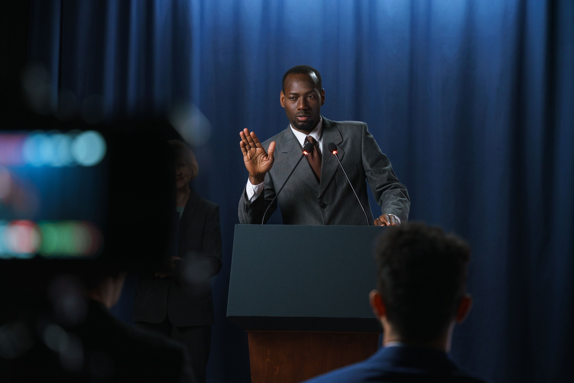 Young serious African-American speaker in a gray suit gesturing during his speech at the debates with a shooting camera in the foreground